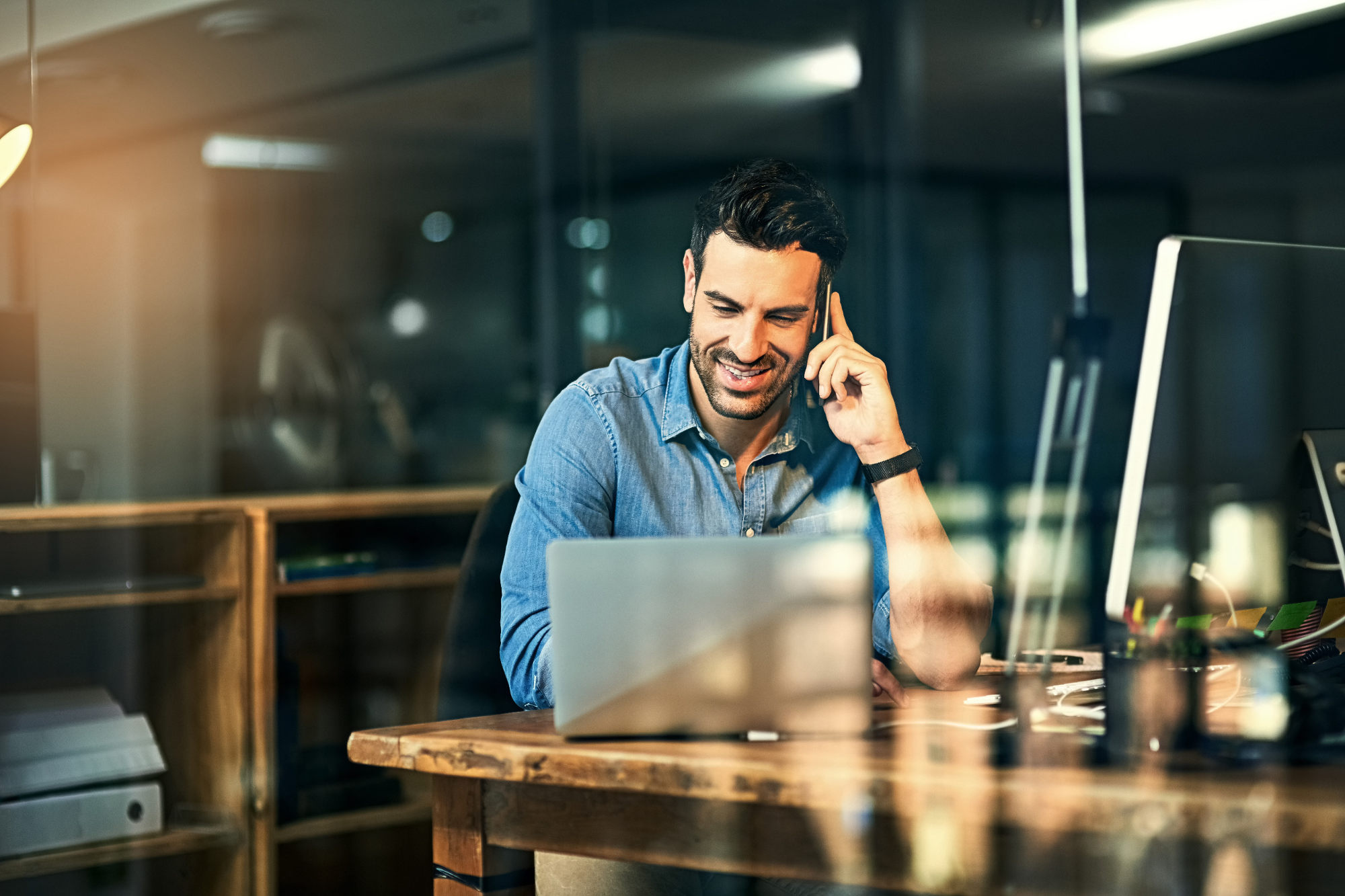 Smiling man talking on smartphone while sitting in front of laptop